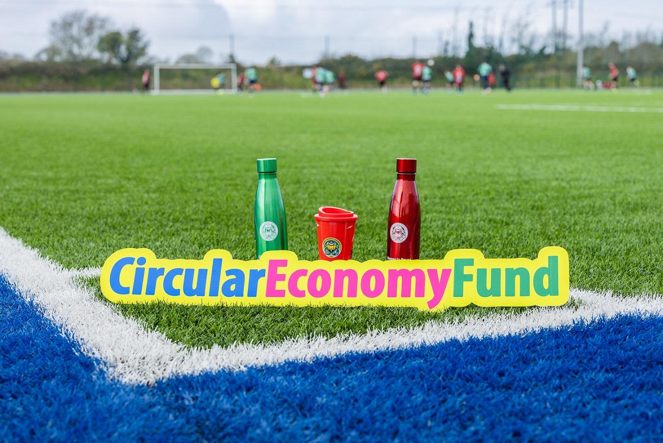 A green reusable metal water bottle, a red reusable metal water bottle and a red resuable coffee mug all with the Cork County logo placed on a sports pitch with the 'Circular Economy Fund' logo placed in front of them.