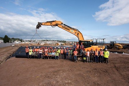 A large group of people in safety vest standing in from of a crane