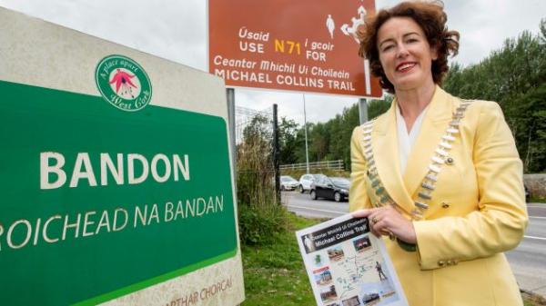 Mayor of the County of Cork, Councilor Gillian Coughlan holding a brochure in front the Michael Collins Trail sign post 