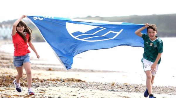 Image of girl and boy running with blue flag on beach. Cork County beaches have been awarded a total of 23 flags by An Taisce at the National Awards for 2021.