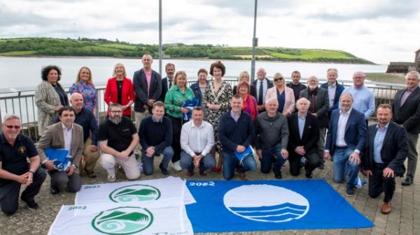 Mayor with Councillors, Community reps and council staff  behind green flags and a blue flag on the ground.