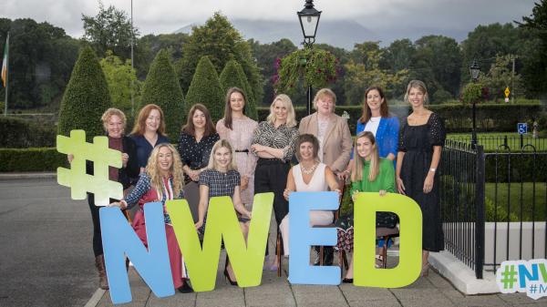 A group photograph of women standing behind and # N W E D sign with trees in the background