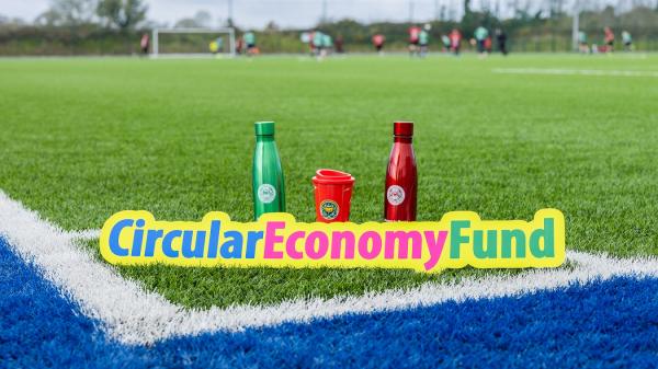 A green reusable metal water bottle, a red reusable metal water bottle and a red resuable coffee mug all with the Cork County logo placed on a sports pitch with the 'Circular Economy Fund' logo placed in front of them.