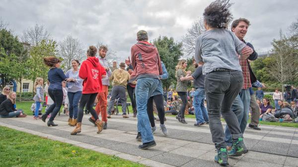 People dancing on a paved path in a park, surrounded by onlookers.