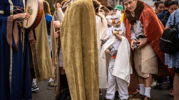 People in medieval-style costumes, including a child in an owl mask and an adult in a red cape, gather outdoors.