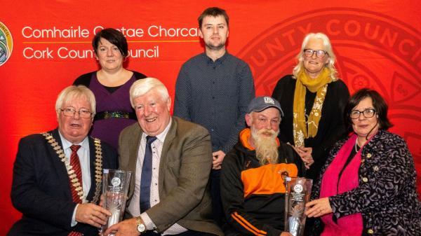 A group of seven people posing with glass awards in front of a Cork County Council backdrop.