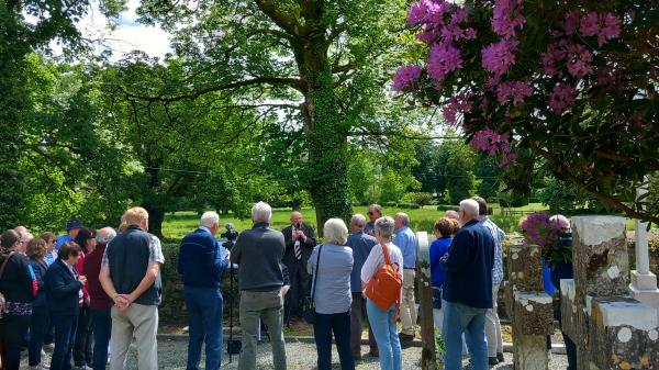 Group of people standing in graveyard underneath trees looking at man speaking into microphone.