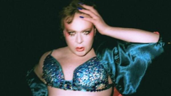 Person wearing a blue sequined top and jacket, hand on head, against a dark background.