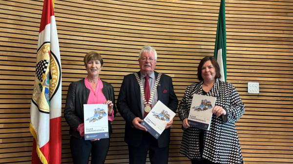 Two women and one man standing in front of a wooden wall between two flags holding a booklet.