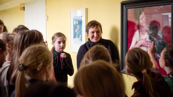 Woman speaking with children next to a painting of a woman with a dog with a red background.