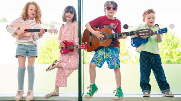  A group of four children holding guitars poses happily in front of a bright window, ready to play music together.