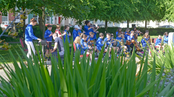 An outdoor musical performance with musicians in blue jackets playing in a park, seen through tall green plants.