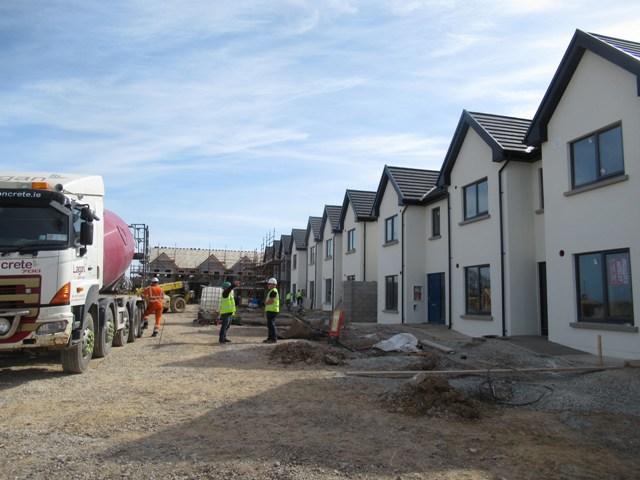 A photograph of works on houses nearly complete with 3 workers and some Construction vehicles operating on the site.