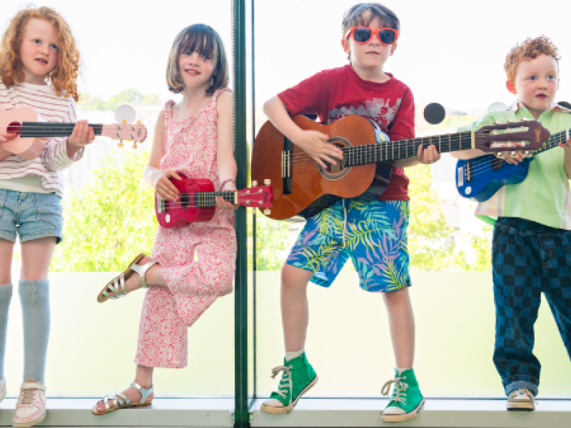  A group of four children holding guitars poses happily in front of a bright window, ready to play music together.