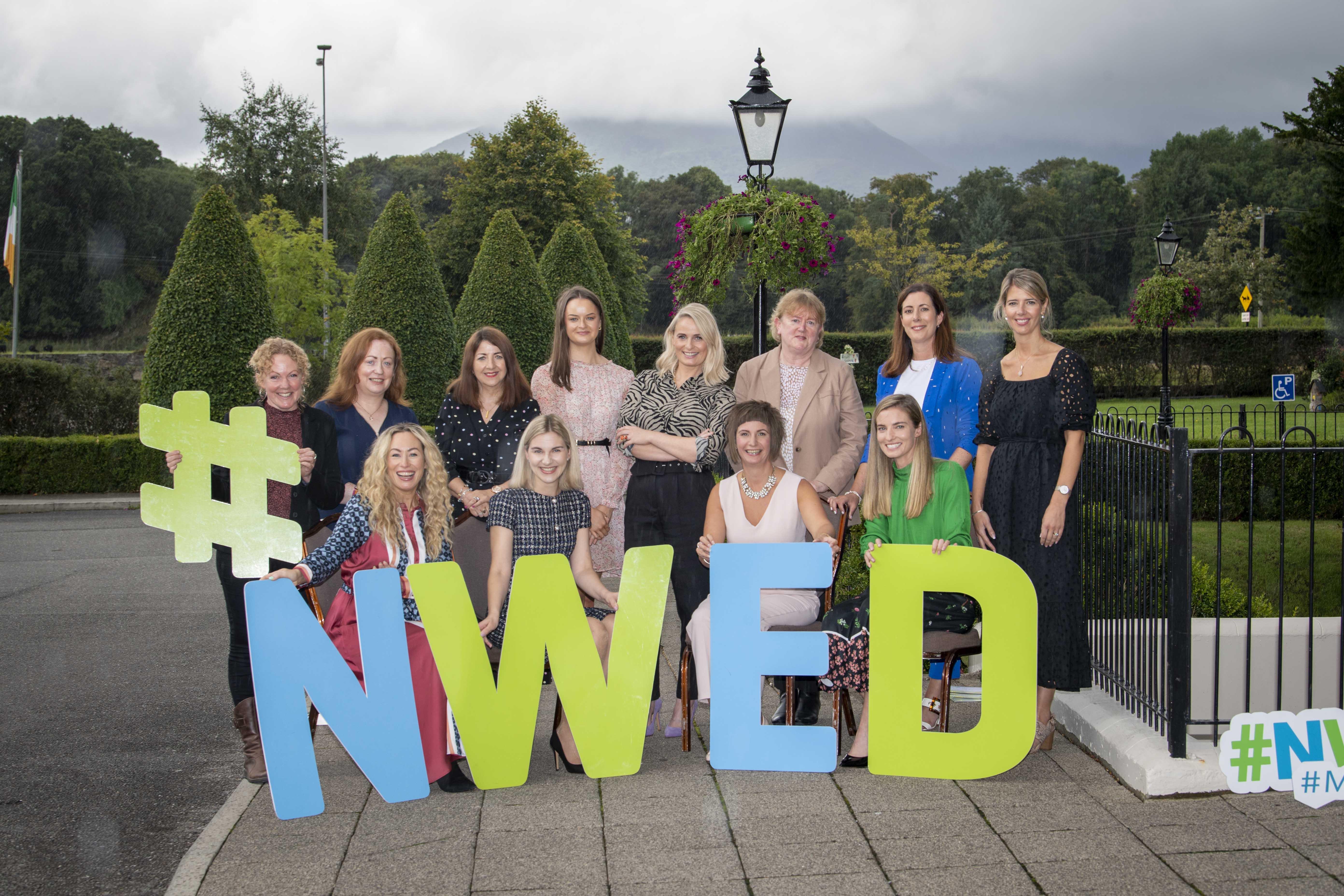 A group photograph of women standing behind and # N W E D sign with trees in the background
