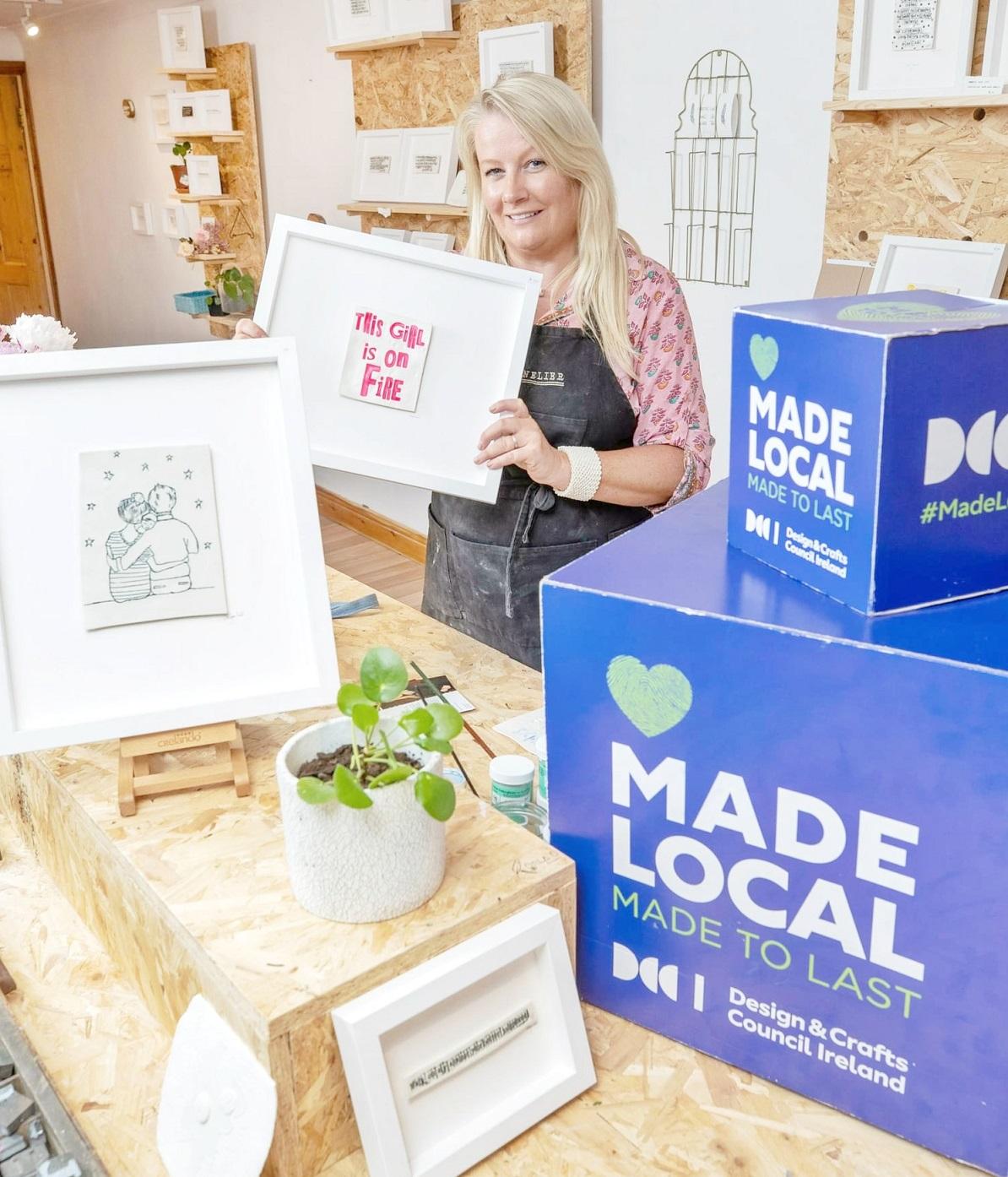 Cork County Council Invites Applications for Creative Startup Scheme 2024. Pictured is a lady holding handmade products alongside a box shaped sign saying 'Made Local, made to last.'