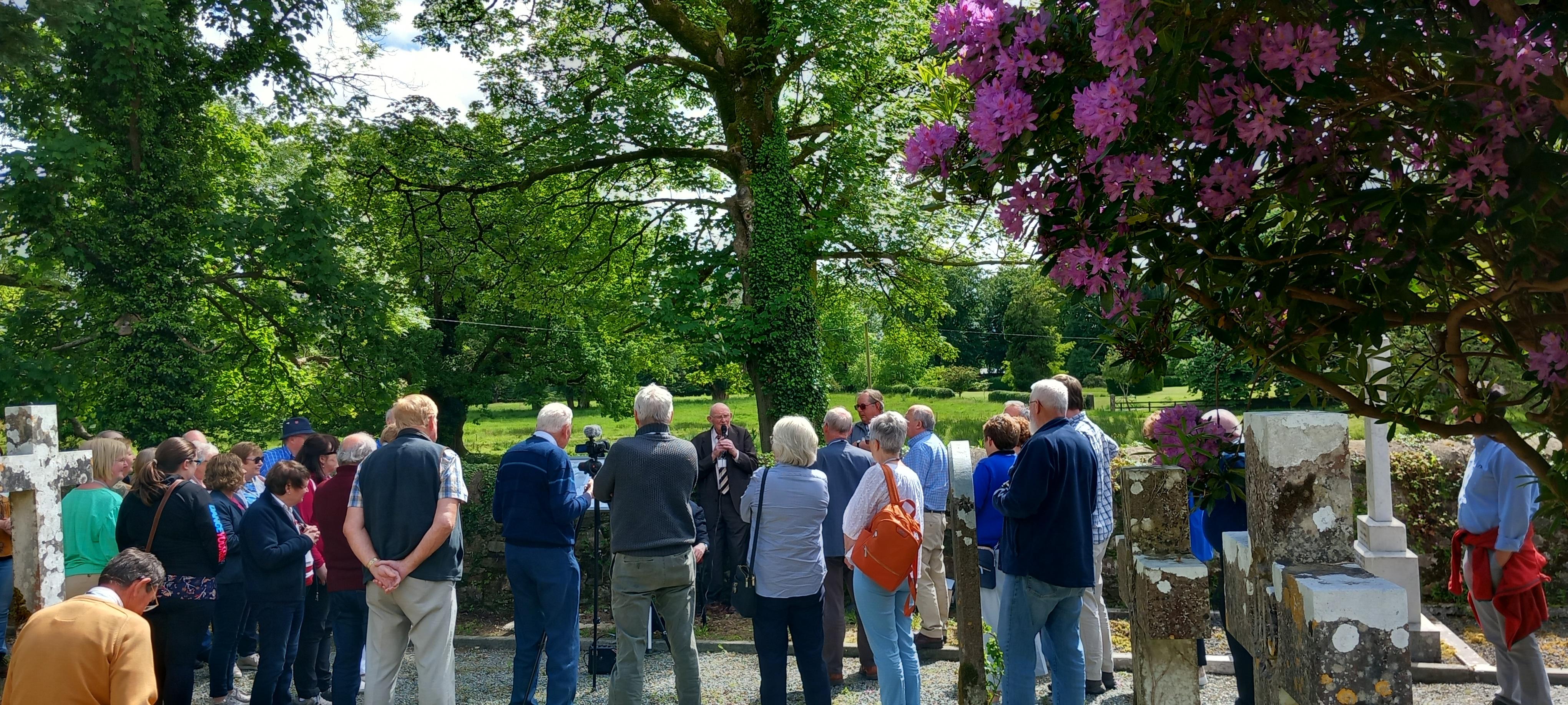 Group of people standing in graveyard underneath trees looking at man speaking into microphone.