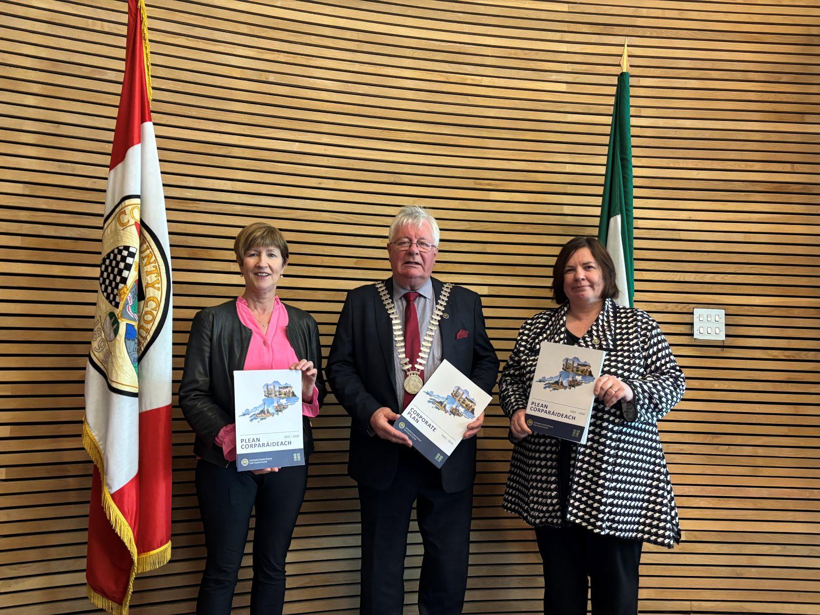 Two women and one man standing in front of a wooden wall between two flags holding a booklet.