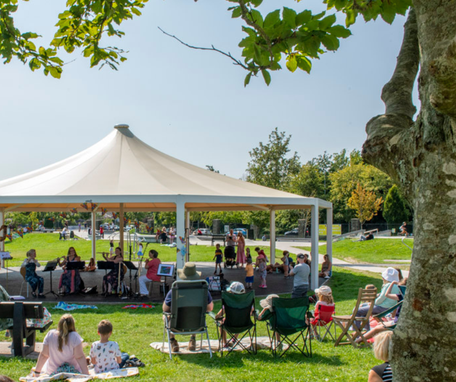 People sitting in a Park on a sunning day listening to musicians play under a Gazebo.