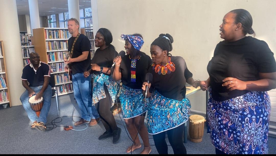Six people in a library singing, dancing and playing instruments inclusing a guitar and drums.
