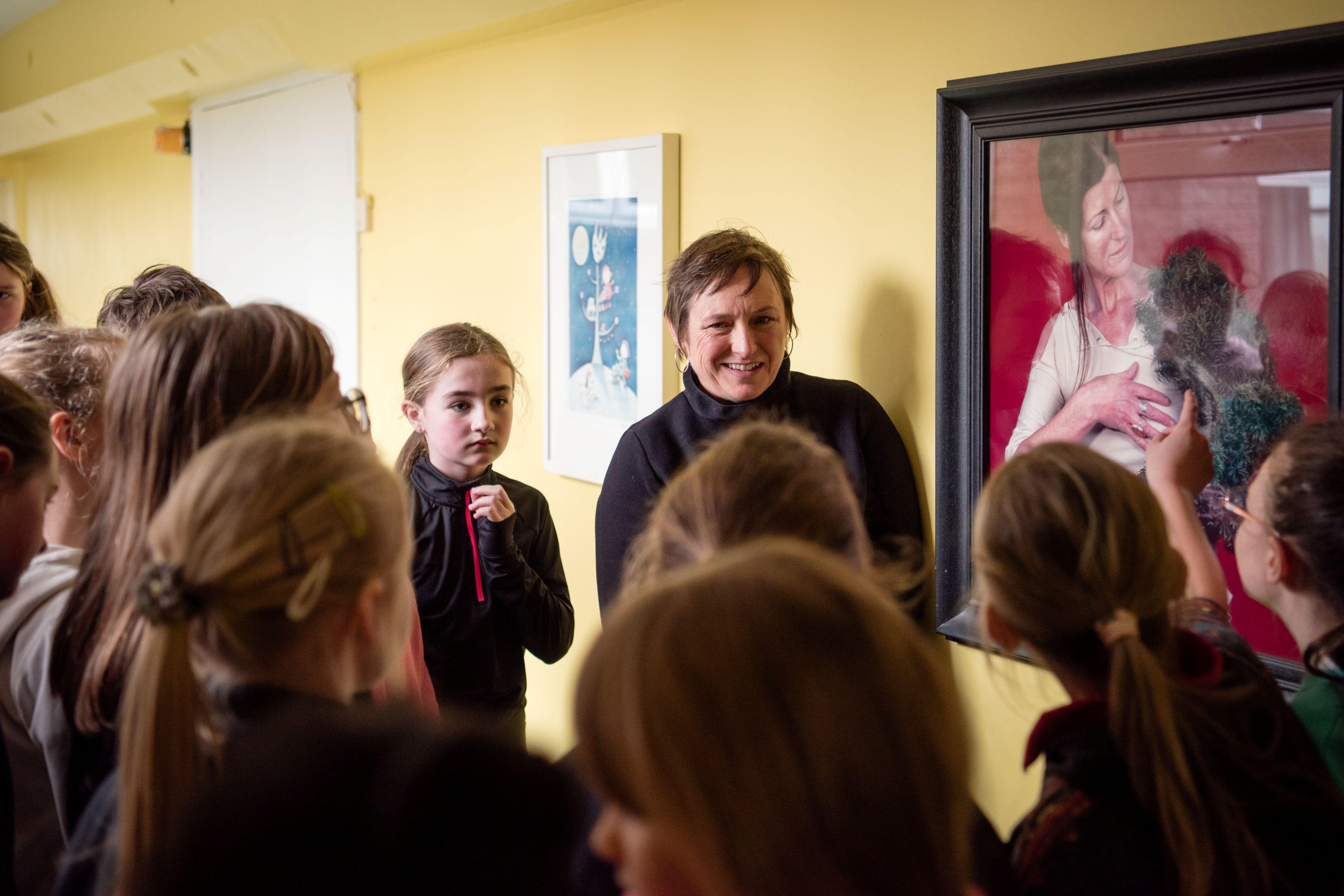 Woman speaking with children next to a painting of a woman with a dog with a red background.