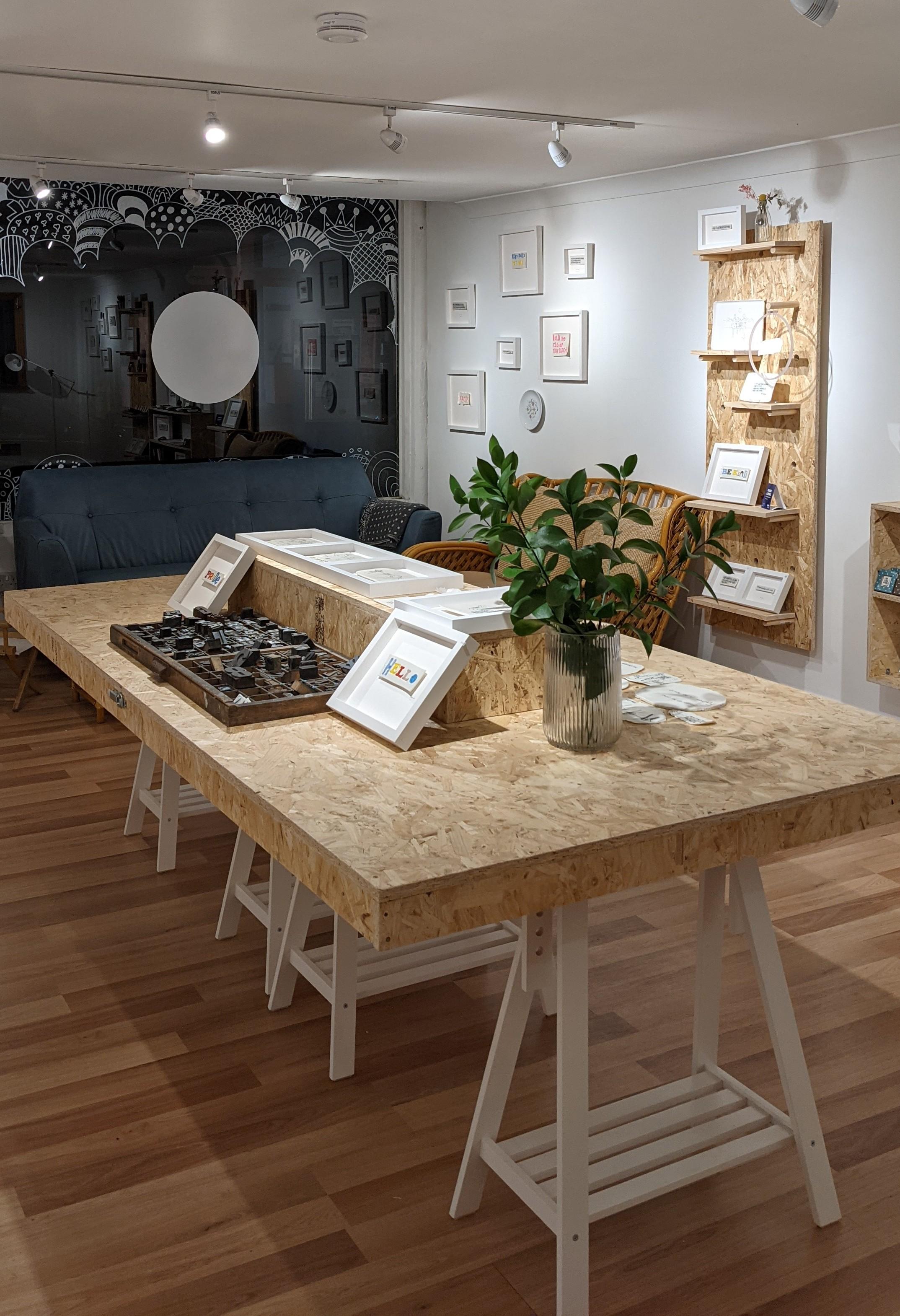 A wooden table on a wooden floor with plants and pictures frames on the table. The walls are black and white and also have pictures.